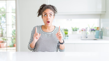 Poster - Beautiful african american woman with afro hair wearing casual striped sweater amazed and surprised looking up and pointing with fingers and raised arms.