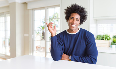 Sticker - Young african american man wearing casual sweater sitting at home with a big smile on face, pointing with hand finger to the side looking at the camera.