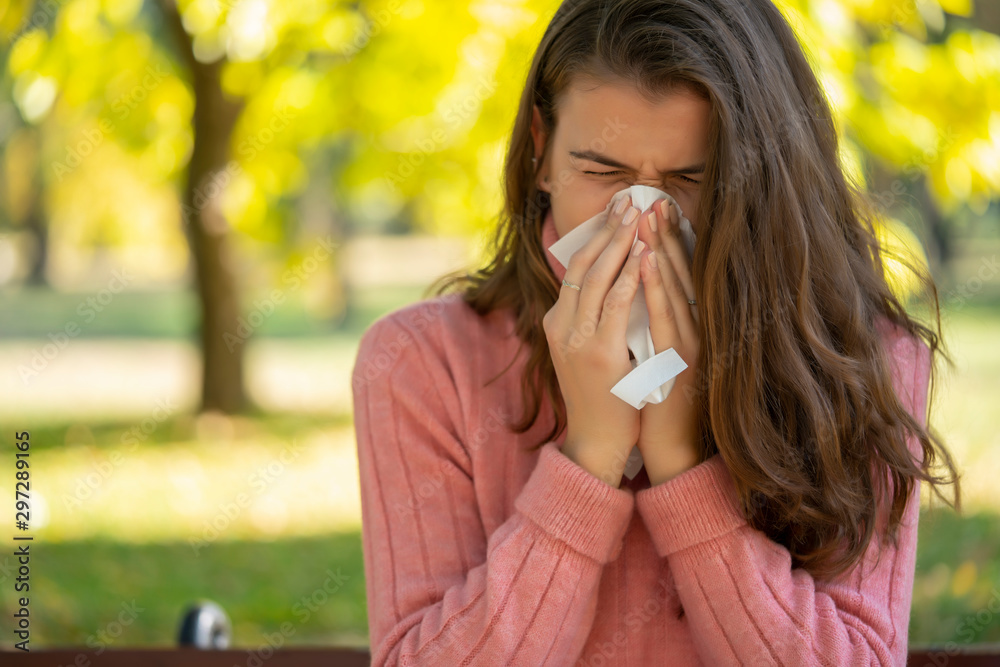 Young woman with allergy sneezing and blowing her nose in a handkerchief tisue, outdoor in a park. - obrazy, fototapety, plakaty 