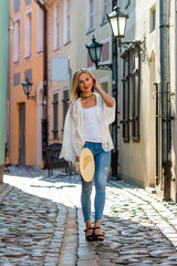 A beautiful woman in a light hat, with long blond hair, a white blouse and blue jeaans in the middle of old town street - image