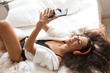 Image of woman wearing headphones and holding smartphone on bed at home