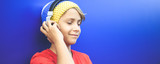 Fototapeta Młodzieżowe - Portrait of a smiling beautiful boy listening music with headphones isolated on a blue background. Colorful image of young male wearing a wool cup, have fun with earphones. Youth fun isolation concept