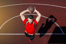 Young Bearded Guy Athlete Performs A Slam Dunk In A Jump. Basketball Player Jumps And Casts A Shadow On The Sports Field. A Beautiful Jump And Toss Of A Basketball Into The Basket.