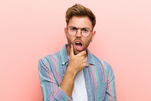 Young Man With Mouth And Eyes Wide Open And Hand On Chin, Feeling Unpleasantly Shocked, Saying What Or Wow Against Pink Background