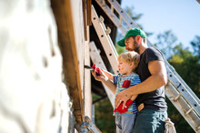 A Father And Toddler Boy Outdoors In Summer, Painting Wooden House.