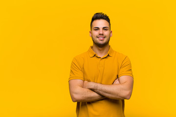 young hispanic man looking like a happy, proud and satisfied achiever smiling with arms crossed against orange wall