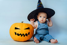 Little Baby In A Big Black Hat Sits Next To An Orange Pumpkin. Halloween Concept