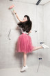 Young woman with pink chiffon skirt and sneakers jumping in front of a gray wall with paint roller in an apartment undergoing renovation