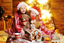 Two Cute Funny Little Girls In Red Santa Hats Covered With A Plaid Smile Against The Background Of Christmas Decor And Lights And Make A Beautiful Gingerbread House
