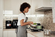 © nenadaksic - Side view of adorable mixed race woman in apron standing next to stove and making lunch.