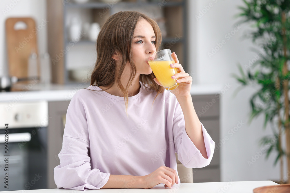 Beautiful young woman drinking orange juice in kitchen - obrazy, fototapety, plakaty 