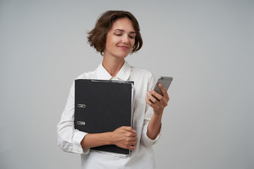 Wall Mural - Portrait of pretty brunette woman with short haircut holding folder with documents and typing message on her mobile phone, being in nice mood, isolated over white background