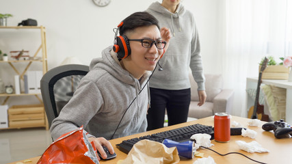 Wall Mural - asian young man in headset playing online video game on computer intense concentrated. unrecognized angry mad annoyed girlfriend talking to him standing beside in dirty apartment. male ignored girl