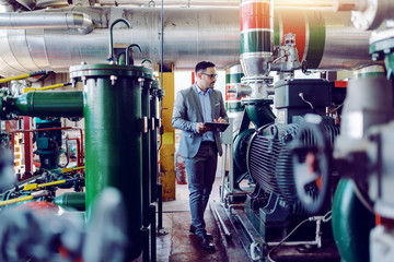 Full length of handsome caucasian supervisor in suit and with helmet on head holding tablet in hands and walking. Power plant interior.