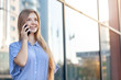 © TravelMedia - Happy smiling attractive young woman with long hair calling on mobile phone outside the office building near the windows. Copy space. Working female concept.