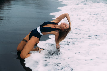 Wall Mural - Sexy girl in blue swimwear sits on beach with black sand and big waves