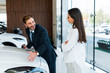 © LIGHTFIELD STUDIOS - happy bearded car dealer gesturing near attractive woman standing with crossed arms in car showroom