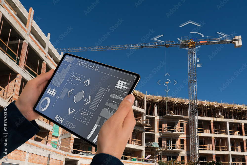 Builder with a digital tablet controls a remote-controlled unmanned crane. Digital transformation in construction industry. - obrazy, fototapety, plakaty 