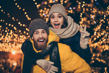 Photo Of Two People Playing Illuminated Night Park X-mas Eve Husband Guy Carry Piggyback Wife Lady Showing V-sign Symbol Wear Winter Jackets Scarfs Hats Outdoors