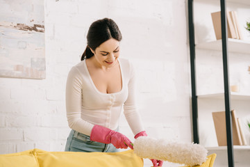 Wall Mural - smiling housewife cleaning yellow sofa with white dusting brush