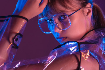 Wall Mural - Fashion sexy young woman girl model attractive face wear stylish trendy transparent raincoat eyewear sunglasses looking at camera in neon light at purple magenta studio background, close up portrait