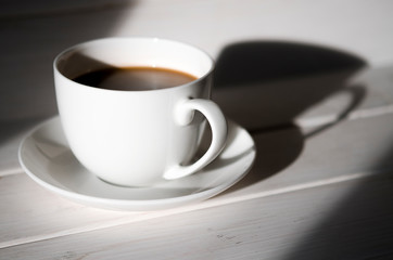 A round white cup of coffee stands on a white saucer on a white wooden table next to the shadow of the cup