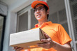 Happy young delivery man in the orange uniform standing with  parcel box to send to customers, focus on the parcel, delivery concept.
