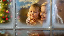 On Christmas Eve Mother And Daughter Looking Through Window. Garland Shines Bright On A Window.
