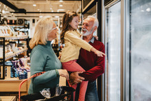Happy Grandmother And Grandfather With Granddaughter Shopping Together In Grocery Store Or Supermarket. Consumerism Concept.