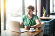 © khosrork - Young satisfied businessman in green t-shirt sitting and working on laptop near window and showing thumbs up like sign. business and freelancing concept. indoor shot near big window at daytime.