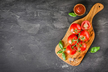 Wall Mural - Fresh red tomatoes on dark olive cut board or dark stone table. Tomato vegetables with ketchup, basil leaves on black background.