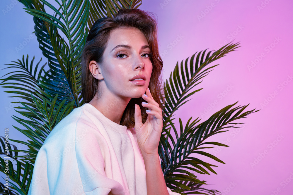 Beautiful female touching face with tenderness. Lady sitting near green palm tree and looking at camera with tenderness. Beauty and fashion concept - obrazy, fototapety, plakaty 