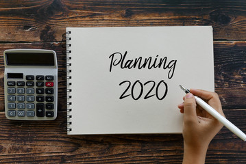 Fototapete - Top view of calculator, and hand holding pen writing Planning 2020 on notebook on wooden background.