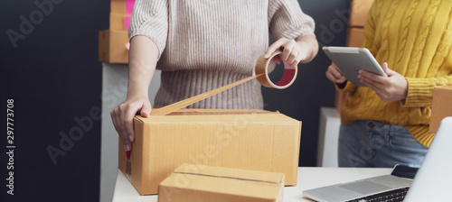 Fotomural Women packing parcels with sticky tape, Entrepreneur owner using smartphone or laptop taking receive and checking online purchase shopping order to preparing pack product box