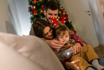 Wall Mural - First Christmas with our cute baby son. Decorating for Christmas holidays. Young couple with a baby standing next to a decorated Christmas tree. Christmas joy