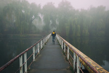 The Man Is Cycling Through The Old Rusty Bridge Over The Lake In The Early Foggy Morning