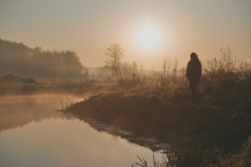 Woman walking through a meadow by a pond in the foggy morning. Sun rising above field and pond flooded with fog in the morning. Real people, authentic situations