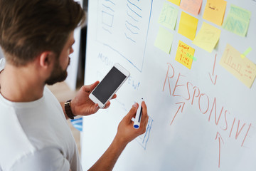 Wall Mural - Web designer looking at smartphone while writing notes on whiteboard in modern office