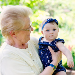 Wall Mural - Cute little baby girl with grandmother on summer day in garden. Happy senior woman holding smiling child on arm..