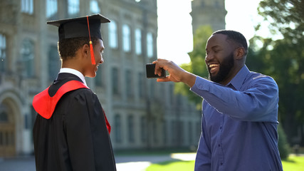 Wall Mural - Happy father taking smartphone photo of glad graduating son with diploma, event