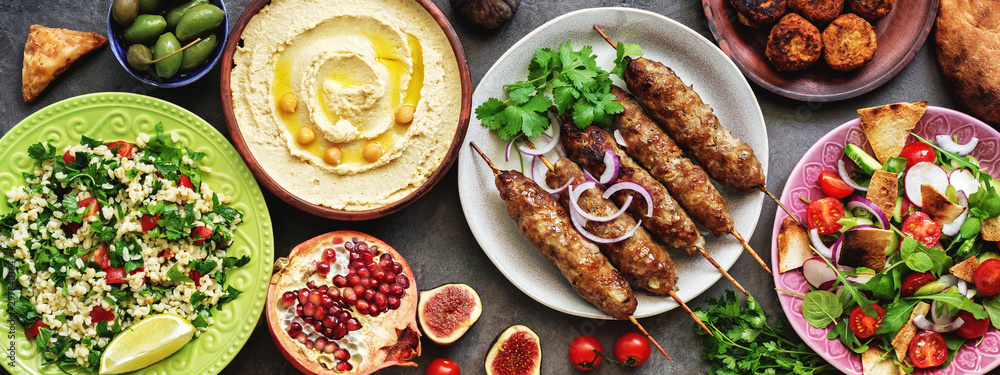 Assorted Middle Eastern and arabic dishes on a dark rustic background,border. Hummus,tabbouleh, salad Fattoush,pita,meat kebab,falafel,baklava,pomegranate. Halal food.Top view, flat lay, copy space - obrazy, fototapety, plakaty 