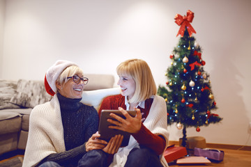 Wall Mural - Happy senior woman with santa hat on head sitting with her daughter on floor in living room and using tablet. Both are covered with blanket. In background is christmas tree.