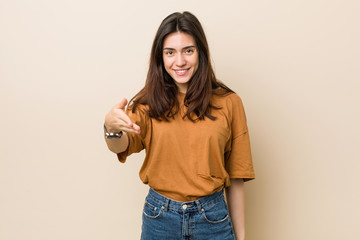 Wall Mural - Young brunette woman against a beige background stretching hand at camera in greeting gesture.