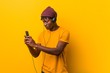 © Asier - Young african man standing against a yellow background wearing a hat listening to music with a phone