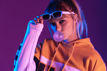 Wall Mural - Stylish pretty young 20s fashion teen girl model wear glasses blowing bubble gum looking at camera standing at purple studio background, igen teenager in trendy night glow 80s 90s concept, portrait