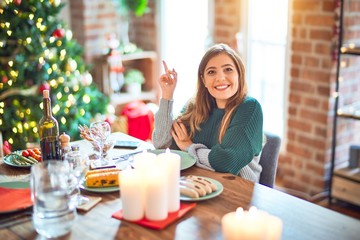 Sticker - Young beautiful woman sitting eating food around christmas tree at home with a big smile on face, pointing with hand and finger to the side looking at the camera.