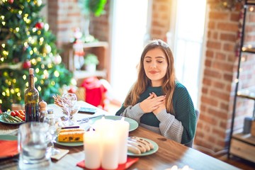 Sticker - Young beautiful woman sitting eating food around christmas tree at home smiling with hands on chest with closed eyes and grateful gesture on face. Health concept.