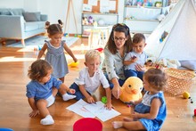 Beautiful Teacher And Group Of Toddlers Sitting On The Floor Drawing Using Paper And Pencil Around Lots Of Toys At Kindergarten