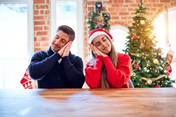 Canvas Print - Young couple wearing santa claus hat sitting on chair and table around christmas tree at home sleeping tired dreaming and posing with hands together while smiling with closed eyes.