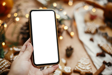 Phone with empty screen in hand on background of christmas lights, festive gingerbread cookies, anise, cinnamon, pine cones, cedar branches on rustic table. Space for text.Merry Christmas